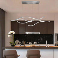 Linear pendant light made of wavy chrome-coloured LED bars hung from a slim rectangular canopy above a kitchen island with a dark stone splashback.