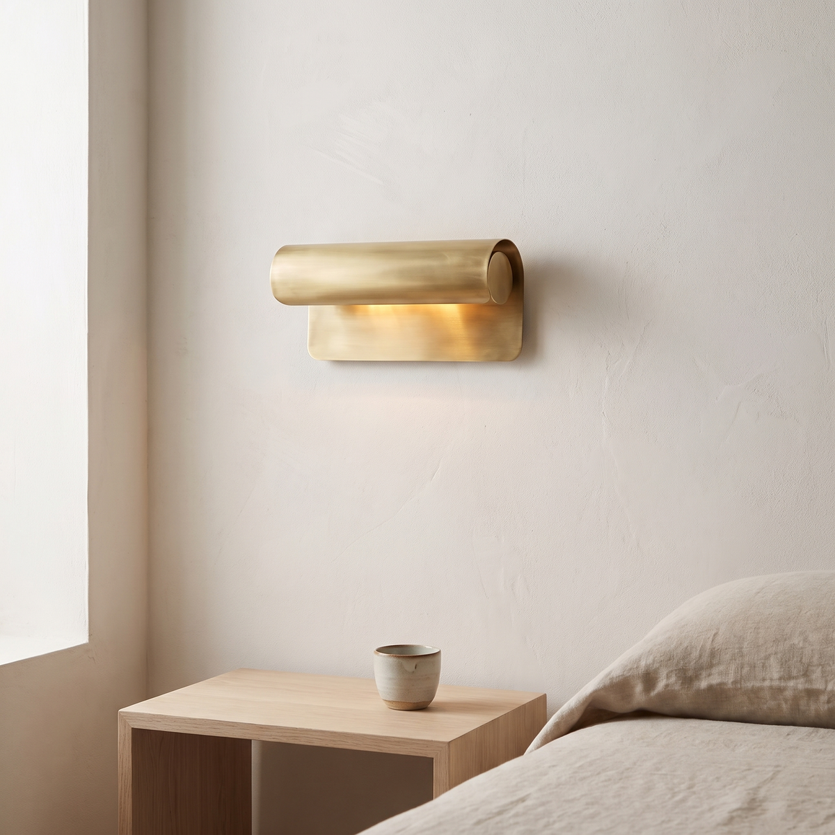 Accelle Wall Light in brushed brass, lit warm on a white plaster wall above an oak bedside table