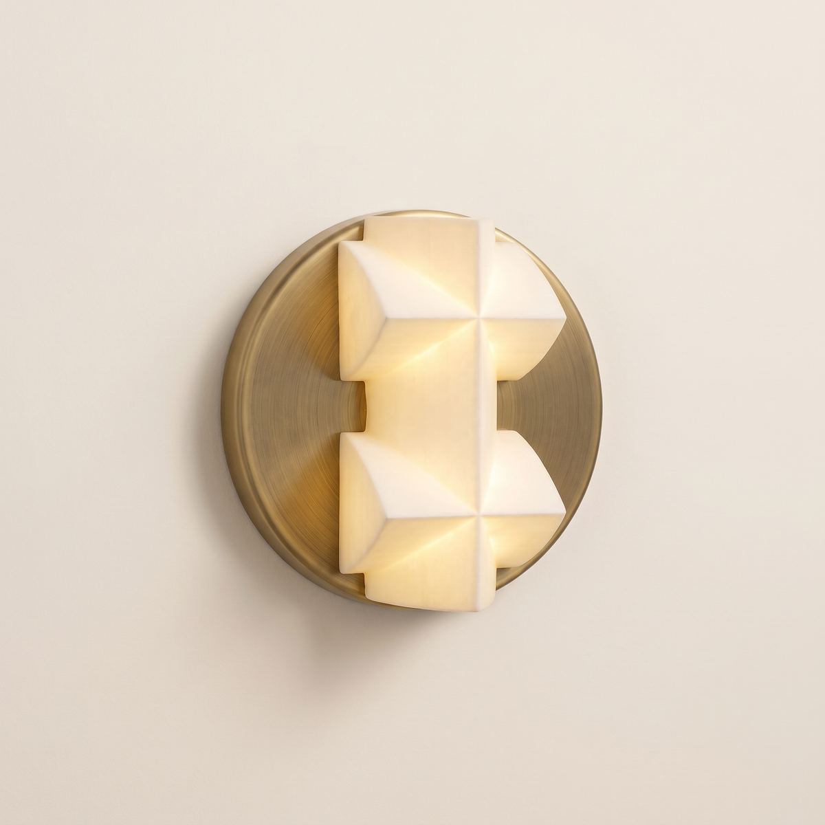 Prismelle Geometric Wall Light: round brushed brass wall plate holding a white faceted geometric alabaster-like block shade shaped like a stepped cross, glowing softly from within