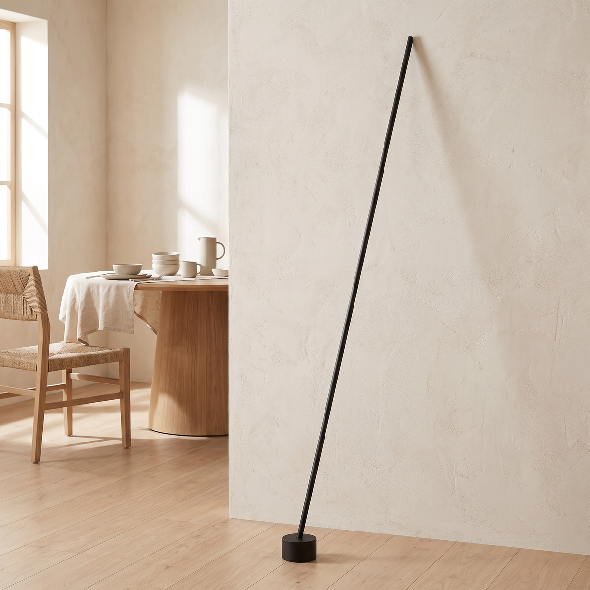 Tensara Elastic Strip Floor Lamp: minimalist floor lamp made of 1 thin straight matte black rod leaning diagonally, its lower end in a small black cylinder floor weight and its top end resting against the wall, shown in a dining room