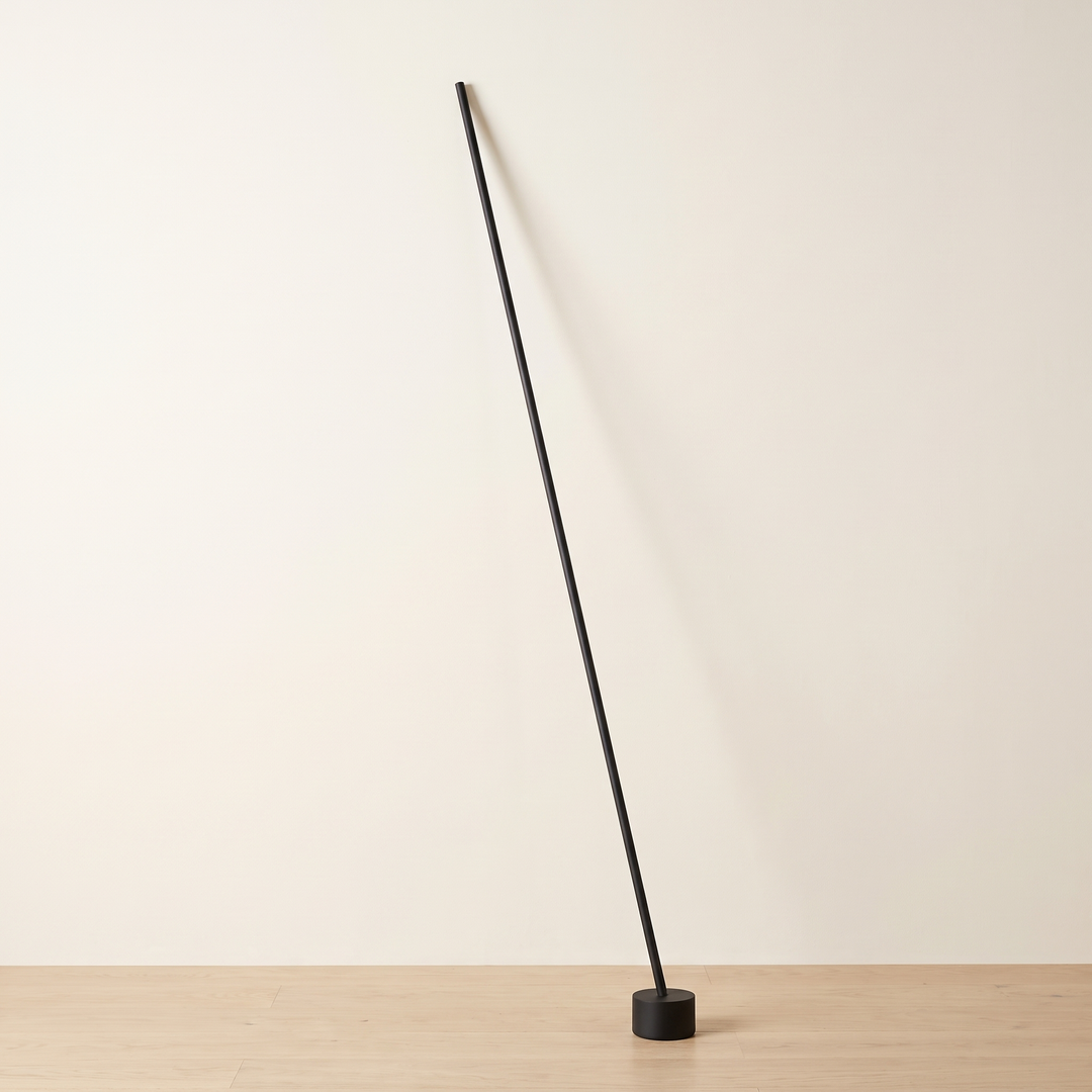 Tensara Elastic Strip Floor Lamp: minimalist floor lamp made of 1 thin straight matte black rod leaning diagonally, its lower end in a small black cylinder floor weight and its top end resting against the wall