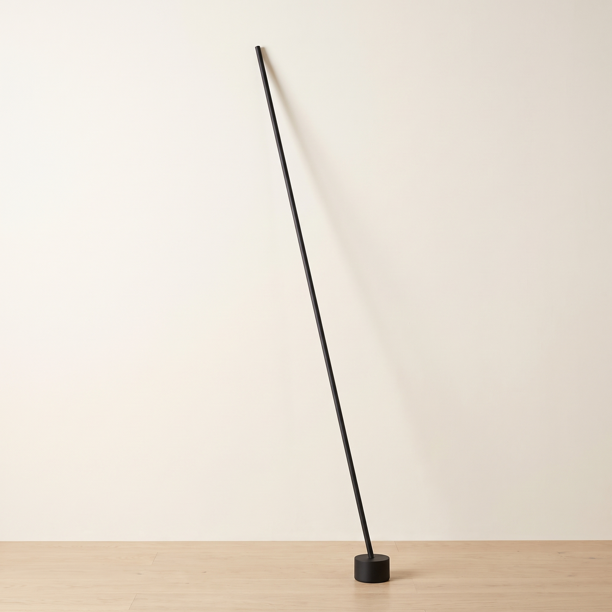 Tensara Elastic Strip Floor Lamp: minimalist floor lamp made of 1 thin straight matte black rod leaning diagonally, its lower end in a small black cylinder floor weight and its top end resting against the wall