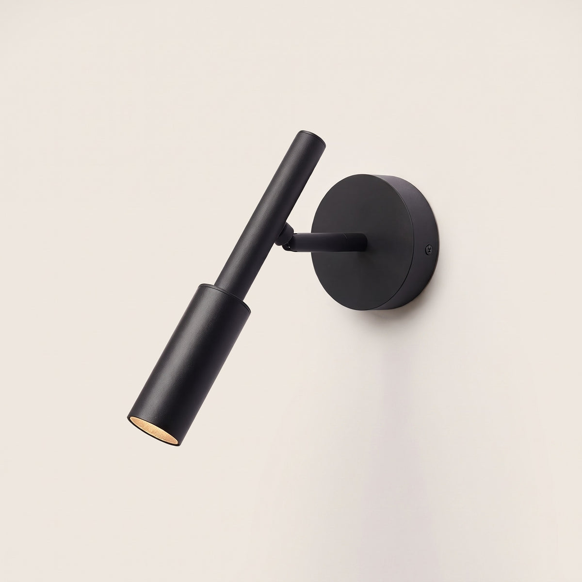 Tuvane Wall Light in matte black with adjustable cylinder head and round wall plate, lit, on a warm off-white studio backdrop