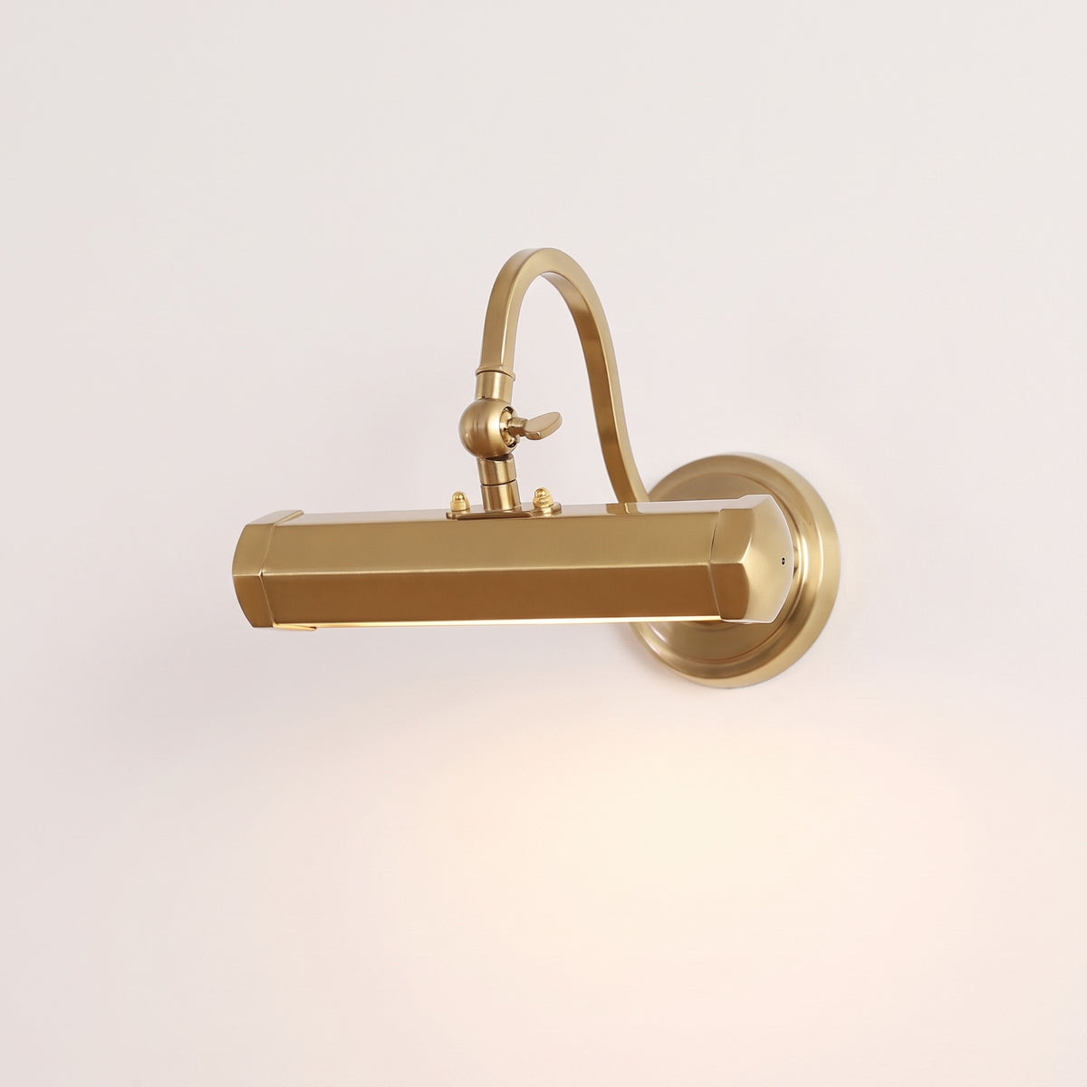 Pacielle Wall Light in gold, a bar picture light on a curved arm with round wall plate, lit, on a warm off-white studio backdrop