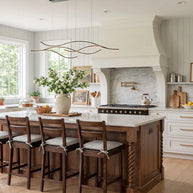 Linear pendant light made of wavy chrome-coloured LED bars hung from a slim rectangular canopy above a kitchen island with wooden stools.