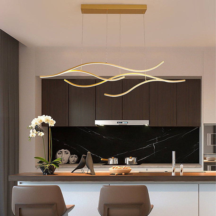 Linear pendant light made of wavy gold-coloured LED bars hung from a slim rectangular canopy above a kitchen island with a dark stone splashback.