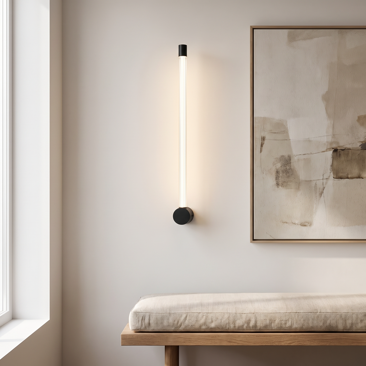 Linear LED Sconce in black, mounted vertically on a pale gallery wall beside a framed canvas, the slim tube reading as a single line of warm light above a bench.