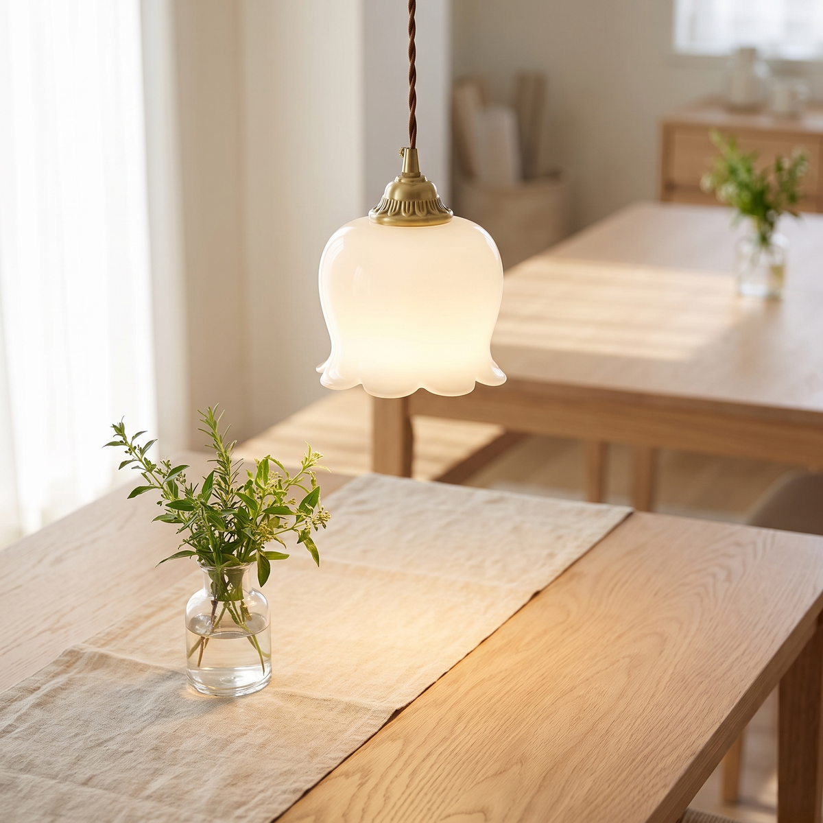 Ravelle pendant with a white bell-flower shade on a brass fitting and twisted cord, hanging above an oak dining table holding a small glass of greenery.