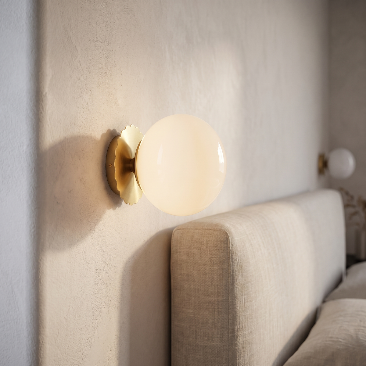 Mirelle wall sconce with a white glass globe on a scalloped gold backplate, mounted on a pale plaster wall beside an upholstered headboard and linen bedding.