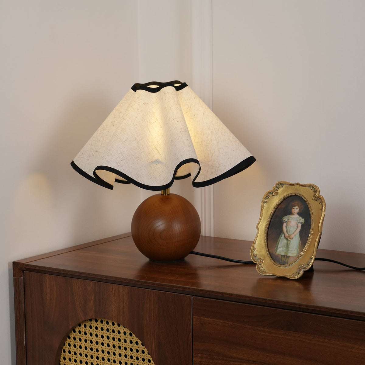 Lit table lamp with a wavy black-trimmed linen shade on a walnut sphere base on a walnut sideboard.