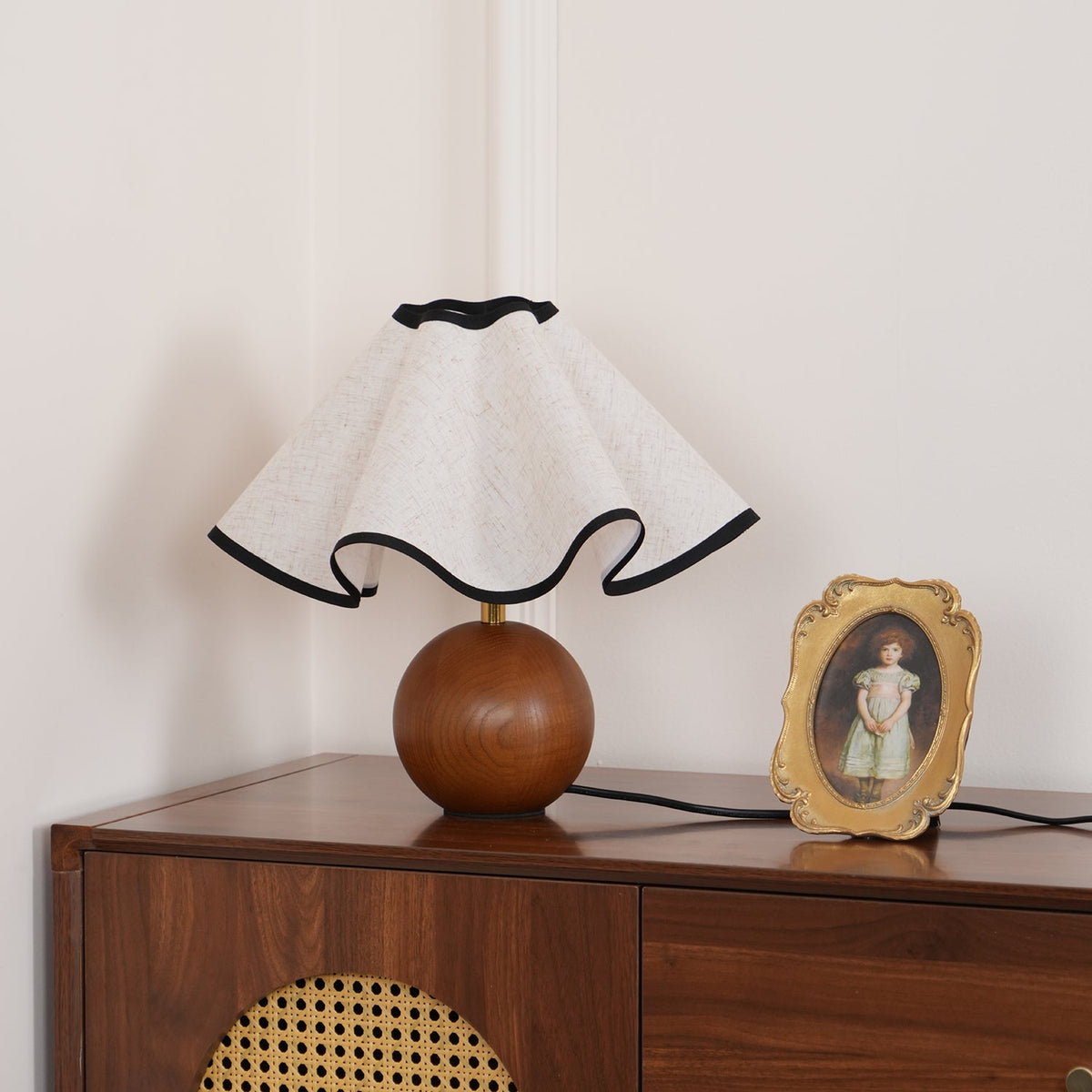 Unlit table lamp with a wavy black-trimmed linen shade on a walnut sphere base on a walnut sideboard.