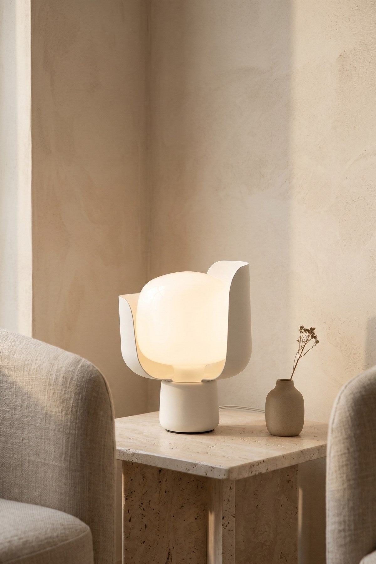 Pethella Table Lamp in white, lit on a stone side table between two armchairs